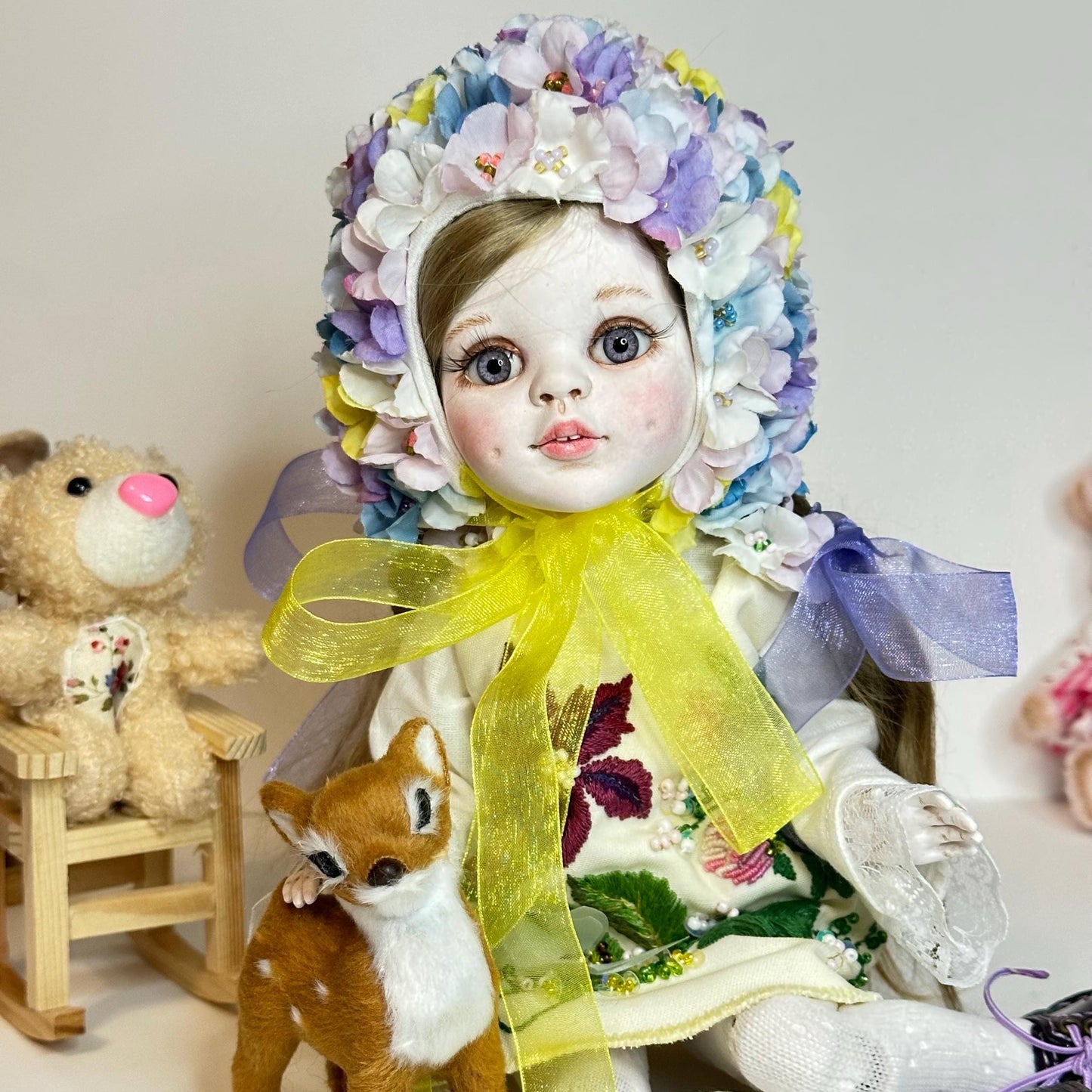 Decorative doll with floral headpiece and yellow scarf, sitting next to a teddy bear and small deer figure.