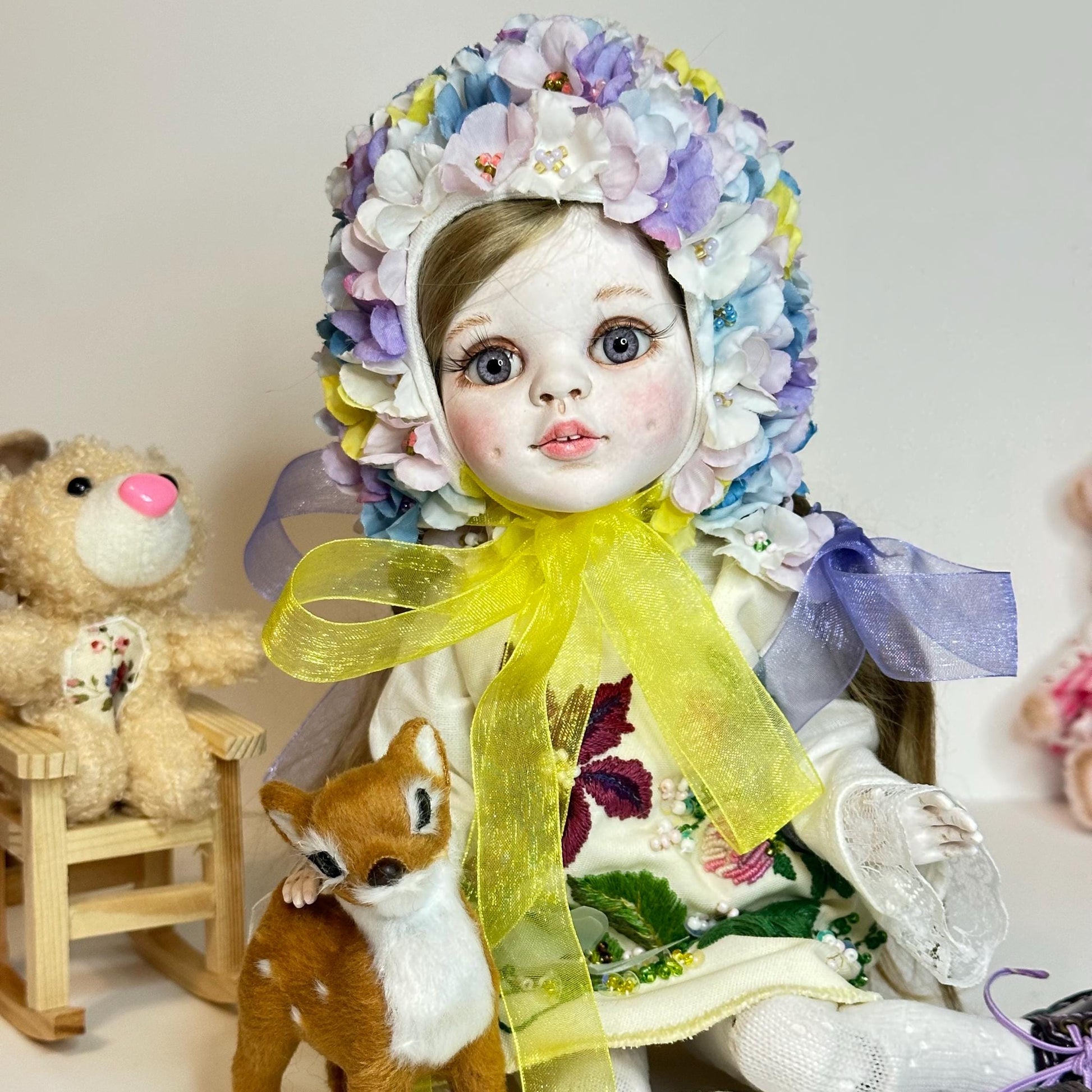 Decorative doll with floral headpiece and yellow scarf, sitting next to a teddy bear and small deer figure.
